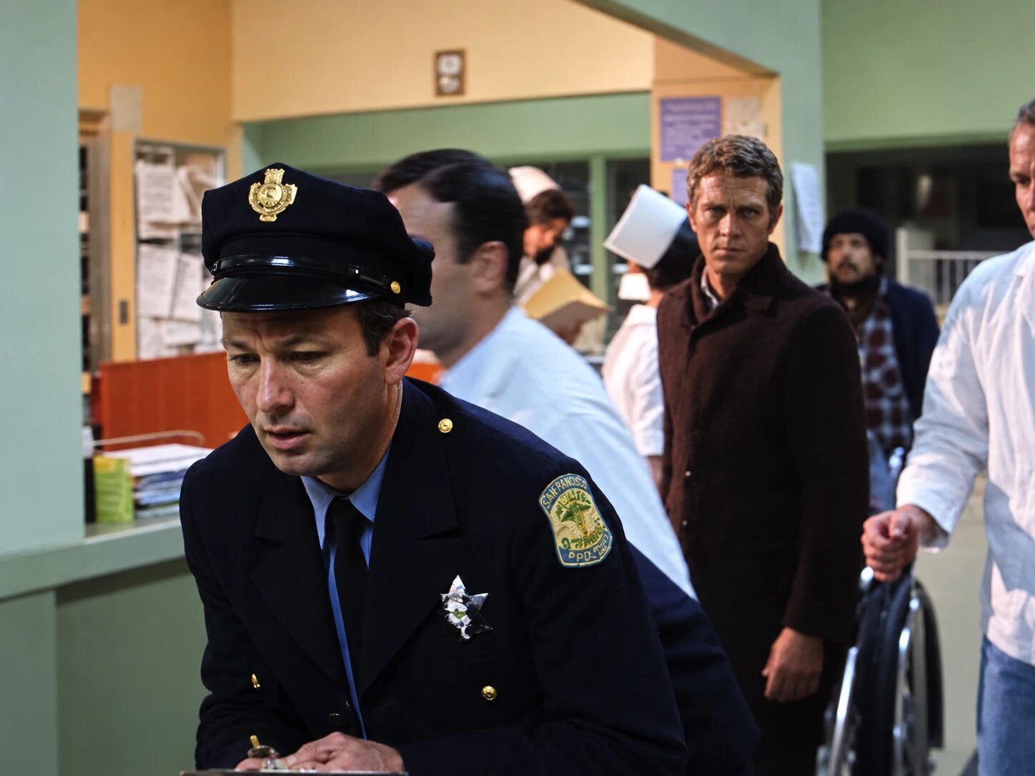 Jim in a San Francisco police uniform writing on a clipboard in a hospital emergency room scene from the 1968 film Bullitt, with Steve McQueen in a brown coat behind him