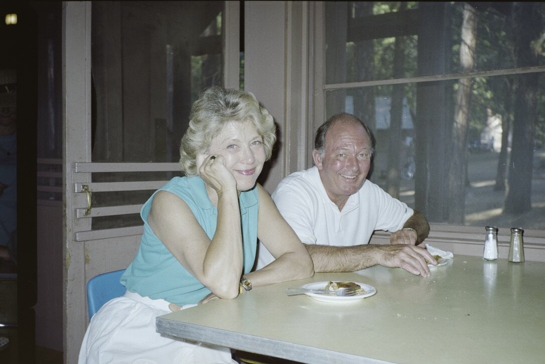 Jan resting her chin on her hand and Jim smiling beside her at a table in the Camp Mather dining hall, tall pines visible through the window