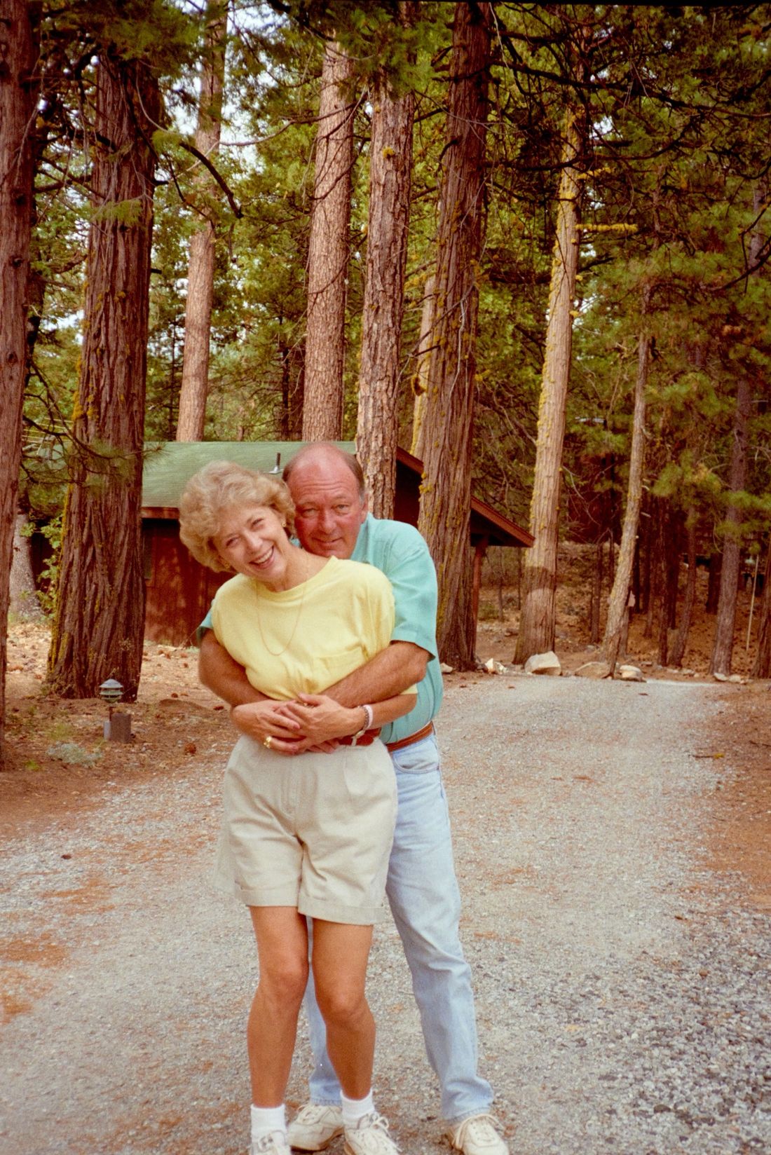 Jim hugging Jan from behind as she laughs, on a gravel road among tall pines at Camp Mather
