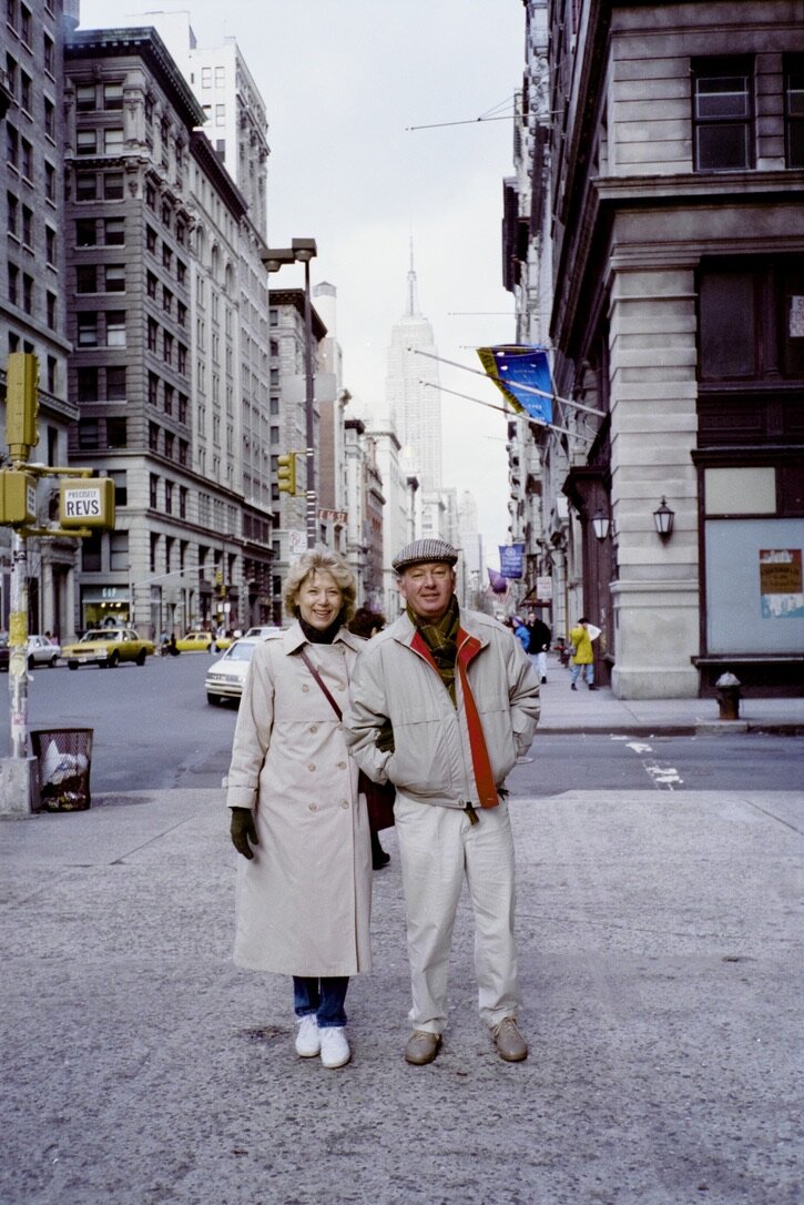 Jim and Jan standing arm in arm on a New York City street corner in winter coats, the Empire State Building rising behind them